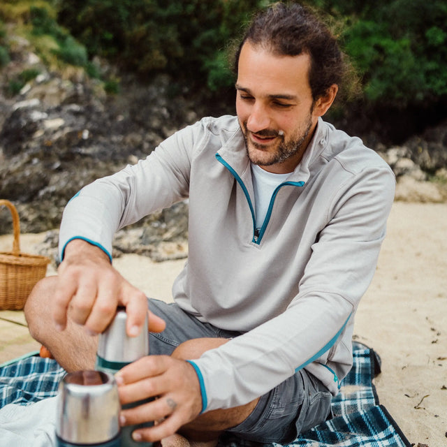 A man enjoying a picnic on the beach wearing a Jellymud Men's Peak Quarter Zip Sweatshirt in grey flint. Made from bamboo and organic cotton.