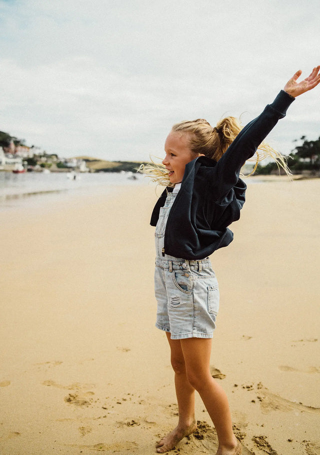 Girl on the beach wearing a Jellymud Juniors' Boulder Zip Through Hoodie in navy.